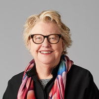 An older lady with short blonde hair smiling at the camera wearing brown glasses and a pink, blue and orange scarf