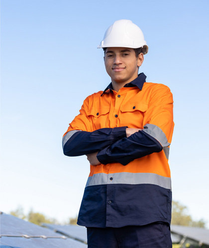 Person wearing a hard hat and high-visibility shirt standing with arms crossed in front of solar panels.