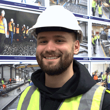 A male student wearing a black hoodie, yellow high vis vest and white hardhat standing in front of a display of construction site visit photographs.