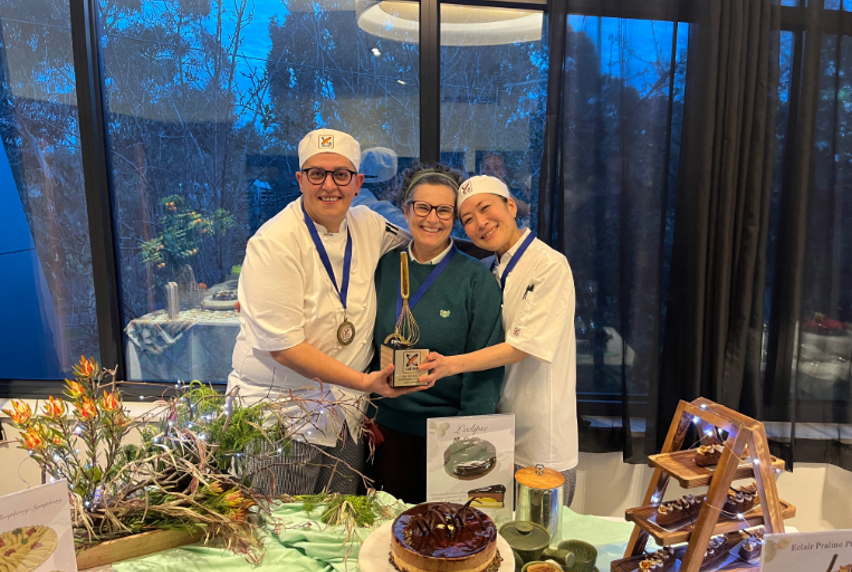 Three people with a whisk-shaped trophy standing behind a table of cakes and éclairs.