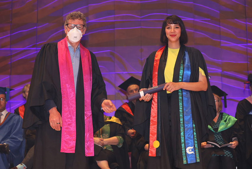 A woman in her graduation gown stands next to a man in a mask, capturing a moment of accomplishment and awareness at her graduation