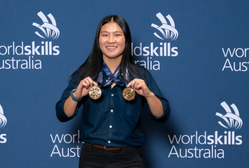 Lily-Grace Toohill dressed in WorldSkills Team uniform in front of a blue media wall, holding her two medals and smiling.