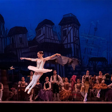 Lead male and female ballerinas on stage dancing with onlooking support dancers in the background 