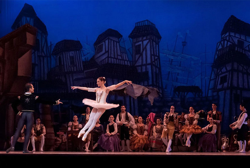 Lead male and female ballerinas on stage dancing with onlooking support dancers in the background