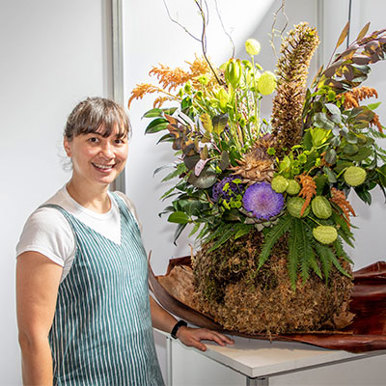 A brunette woman stands beside a sizable flower arrangement smiling