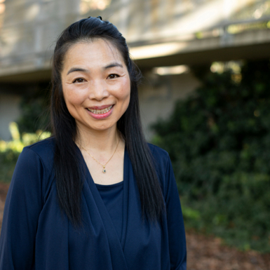  A woman with long black hair wearing a blue shirt, standing and smiling at the camera.