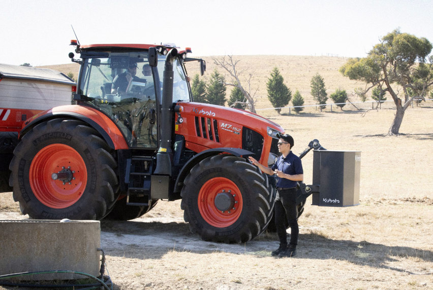 A man standing confidently beside an orange and black Kubota tractor on a property talking to someone off screen