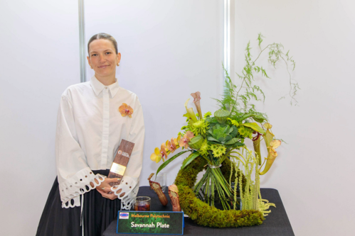 Savannah Plate standing with her floristry arrangement which is made of a green moss structure and spiralled bouquet in green tones.