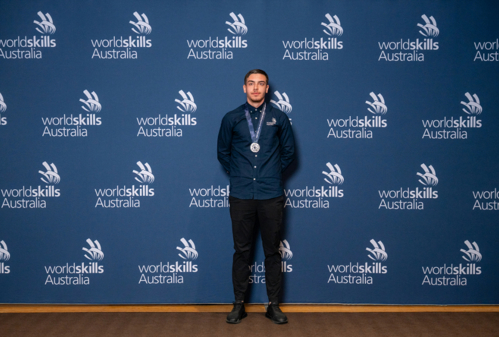 Arif Kamat wearing WorldSkills uniform standing in front of blue media wall with silver medal.