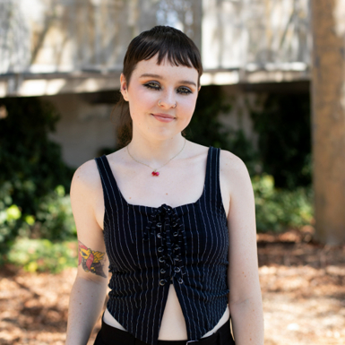 A woman with short brown hair and tattoos, wearing a black top stands in a park, surrounded by greenery and trees.