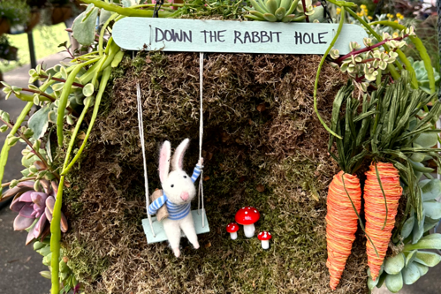 A hanging basket planted with succulent plants and decorated like a rabbit burrow, a sign says 'Down the Rabbit Hole'.