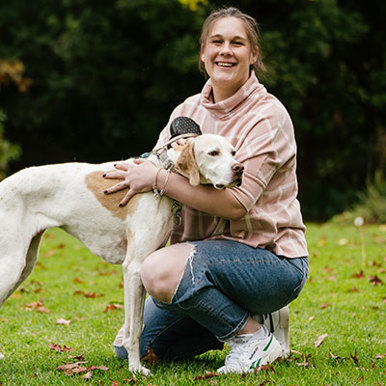 A smiling female wearing a light pink sweater with jeans is kneeing down patting a white spotted dog.