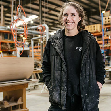 Image of a smiling female, wearing a black vest and jumper covered in dust. She is standing in a timber warehouse.