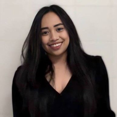 Portrait of a young woman with shoulder-length black hair and a friendly smile, wearing a dark V-neck top. She stands against a plain, light-colored background.