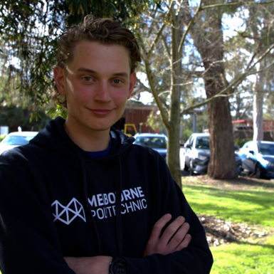 A male student wearing a navy hoodie with Melbourne Polytechnic logo in white, standing with arms crossed in front of a background of trees, lawns and campus carpark.