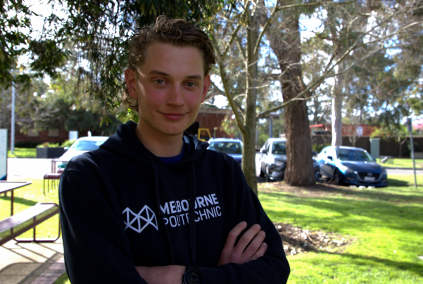 A male student wearing a navy hoodie with Melbourne Polytechnic logo in white, standing with arms crossed in front of a background of trees, lawns and campus carpark.