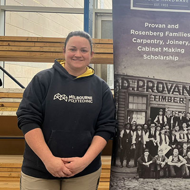 A woman smiling stands beside a banner, wears a dark blue Melbourne Polytechnic jumper.