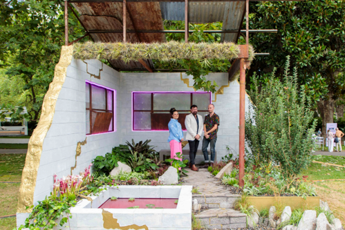 Carla, Alistair and Michael sitting on a grey concrete bench in their garden The New Nature. There is grey paving, green plants and a window behind them surrounded by hot pink neon lights. A roof overhead is rusted and there are overgrown plants growing through the ceiling beams.