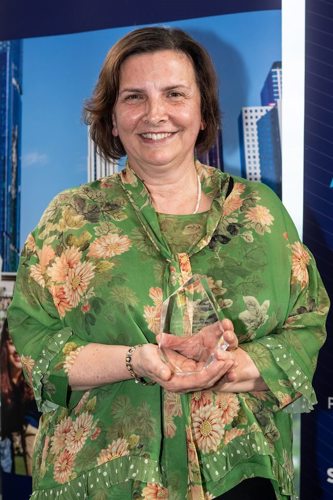 Woman smiling, holding an award, and wearing a bright green floral patterned top.