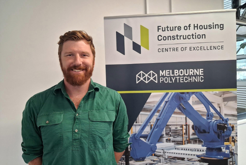 Man smiling in a green shirt stands beside a sign for Melbourne Polytechnic's Future of Housing Construction Centre of Excellence. Background shows industrial machinery.