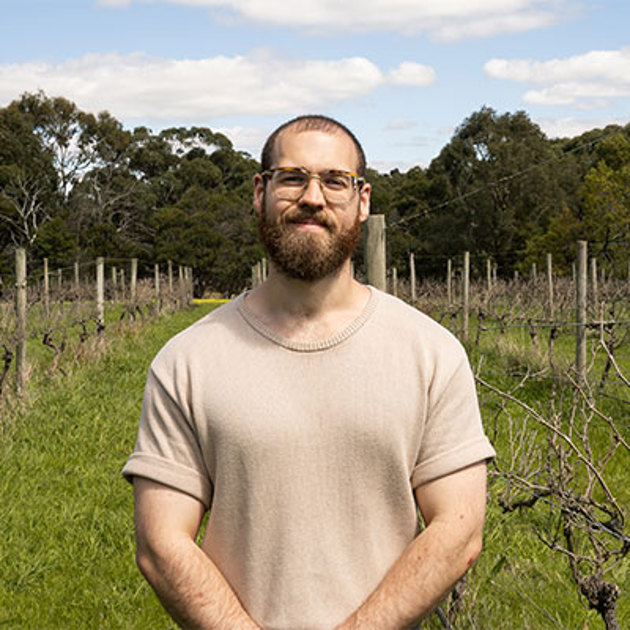 A young man wearing a beige t-shirt stands in a vineyard smiling at the camera