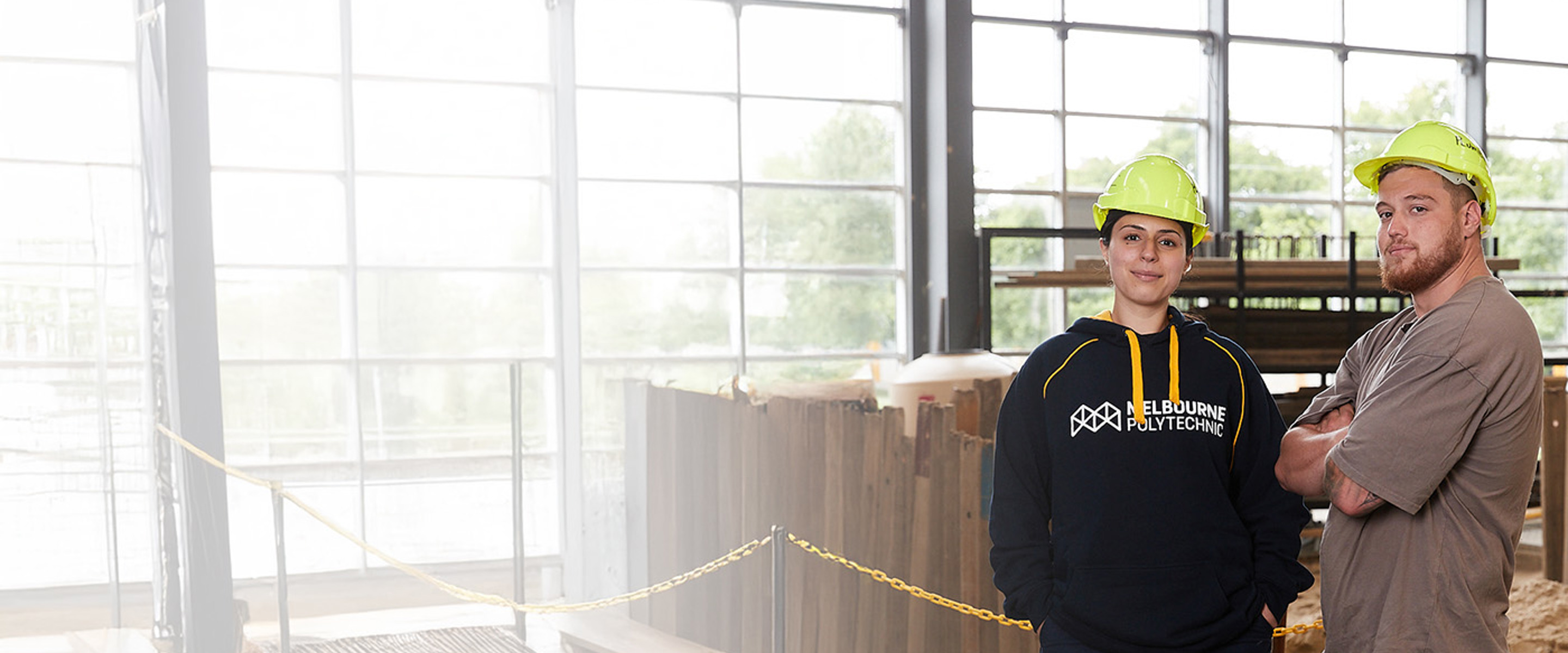 Woman and man posing confidently in a construction workshop wearing hardhats.