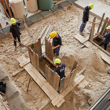 Six plumbing students working on some piping on sandy ground, using tools and equipment to complete the project.