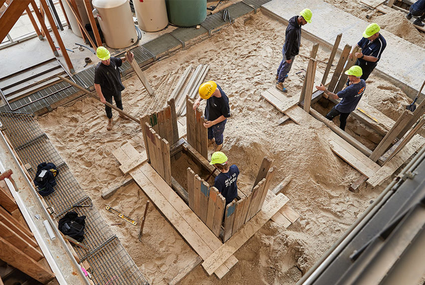 Six plumbing students working on some piping on sandy ground, using tools and equipment to complete the project.