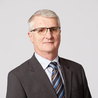 An older man with grey hair, wearing a grey blazer and a brown, blue and navy striped tie, with glasses smiles at the camera.