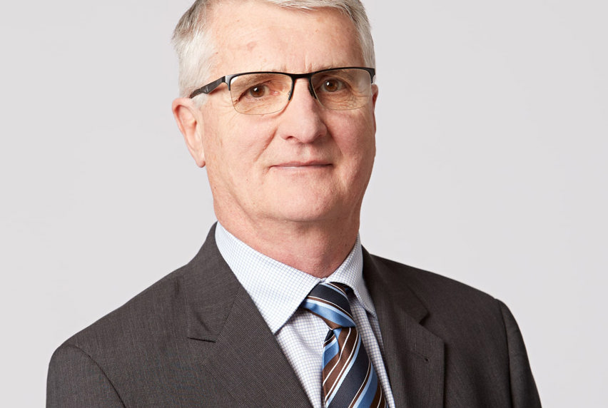 An older man with grey hair, wearing a grey blazer and a brown, blue and navy striped tie, with glasses smiles at the camera.