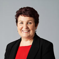 A lady with short dark brown hair, wearing a red top and black blazer smiles at the camera in front of a grey background 