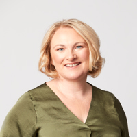 A woman with blonde hair, wearing a green shirt smiles confidently at the camera