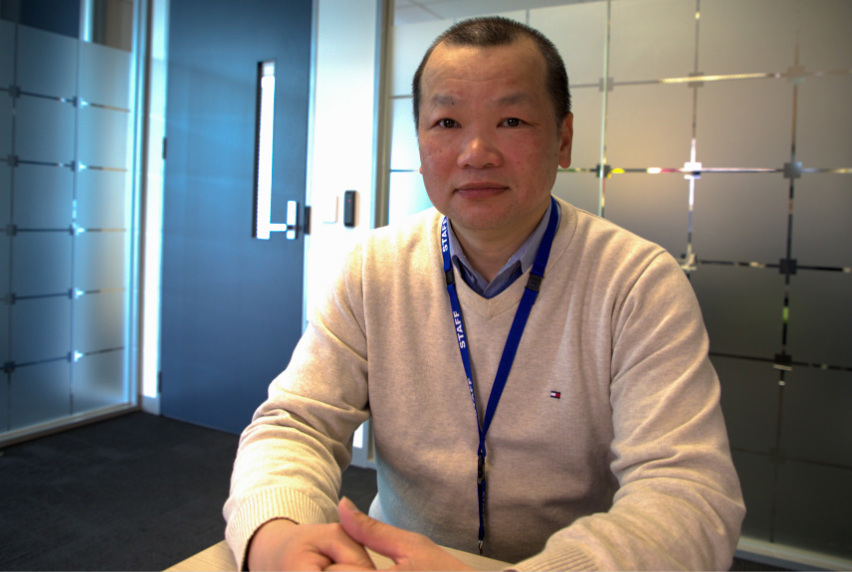 A male student wearing a light blue shirt, cream jumper and a blue lanyard sitting in a meeting room with their hands clasped on the table in front of them.