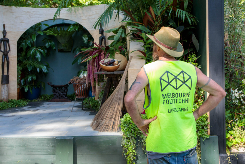 A man dressed in a yellow vest with the Melbourne Polytechnic logo on the back, stands before an award winning garden, gazing at its beauty.
