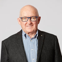 An older man wearing a white and blue stripped shirt, with a charcoal colour blazer and blue glasses smiles at the camera.