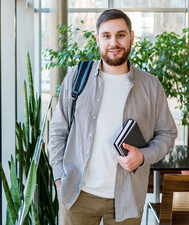 Student standing indoors with backpack and notebooks, smiling at the camera.