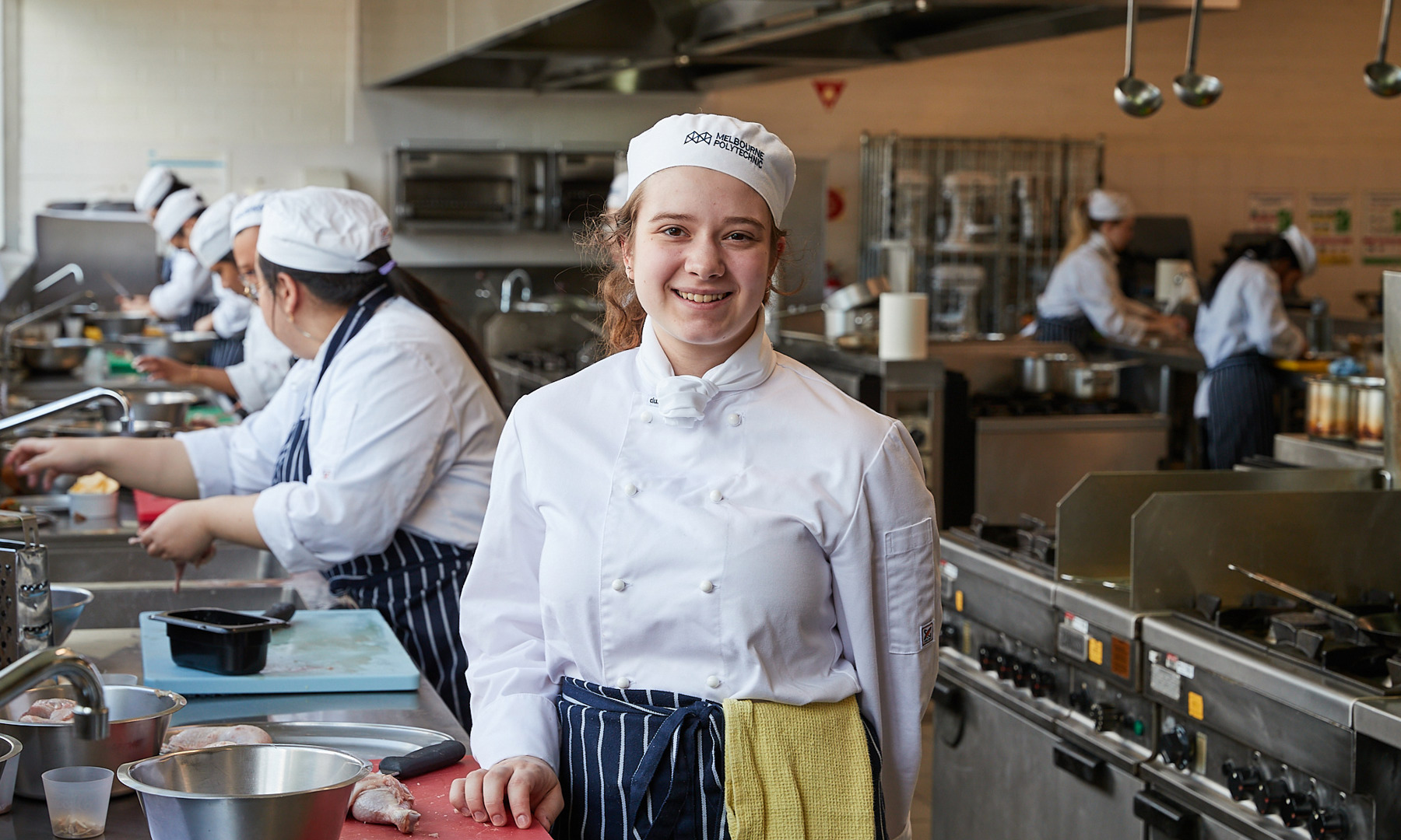 Cookery student in Melbourne Polytechnic cap smiling at the camera during a busy kitchen service with others preparing food in the background.