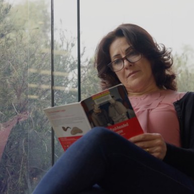 Ekhlas who is a woman, sitting in front of a window with a view of trees, wearing glasses and immersed in reading a book from the library, she is wearing jeans, a pink blouse and black cardigan.