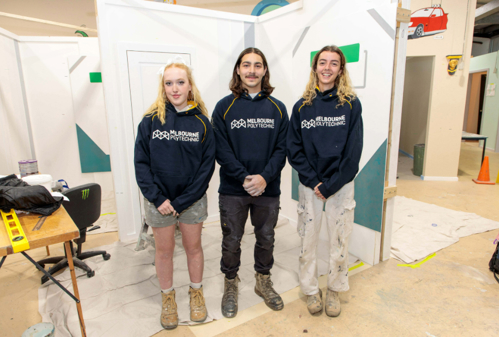 Three Melbourne Polytechnic students wearing hoodies standing in front of competition painting and decorating work area.