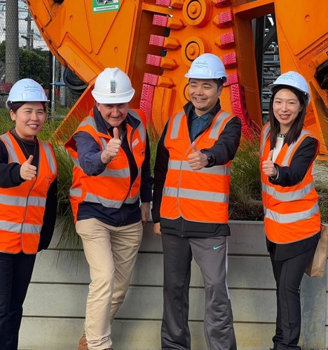 Four people in orange high visibility vests and hard hats standing next to a large orange construction machine.