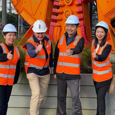 Four people in orange high vis vests and hard hats standing next to a large orange construction machine.