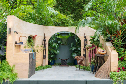 A lush garden planted primarily with tropical plants in green and burgundy tones.  There is a stone tiled floor and rammed earth walls surrounding a kitchen workspace.