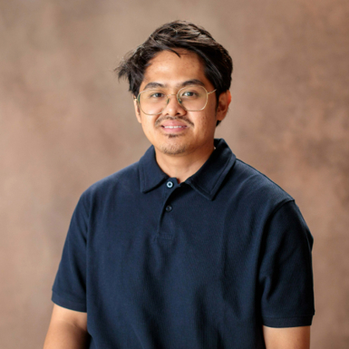 Kino, who is a male IT graduate wearing a navy polo top, glasses looking at the camera and smiling and sitting in front of a beige photography background.