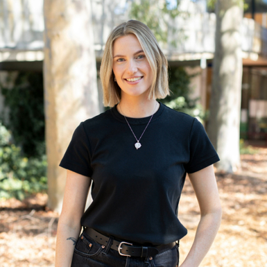 Hannah, an architecture graduate. She is a blonde women with medium-length hair dressed in black tshirt and jeans, stands in a sunlit walkway, smiling. There are trees, shrubs and a brick building in the background.